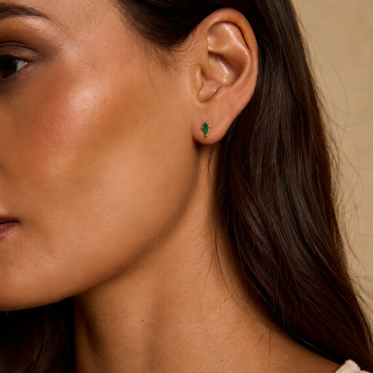 Woman wearing kite cut lab created emerald gemstone stud earrings in 14K yellow gold against a neutral background by Camille Jewelry