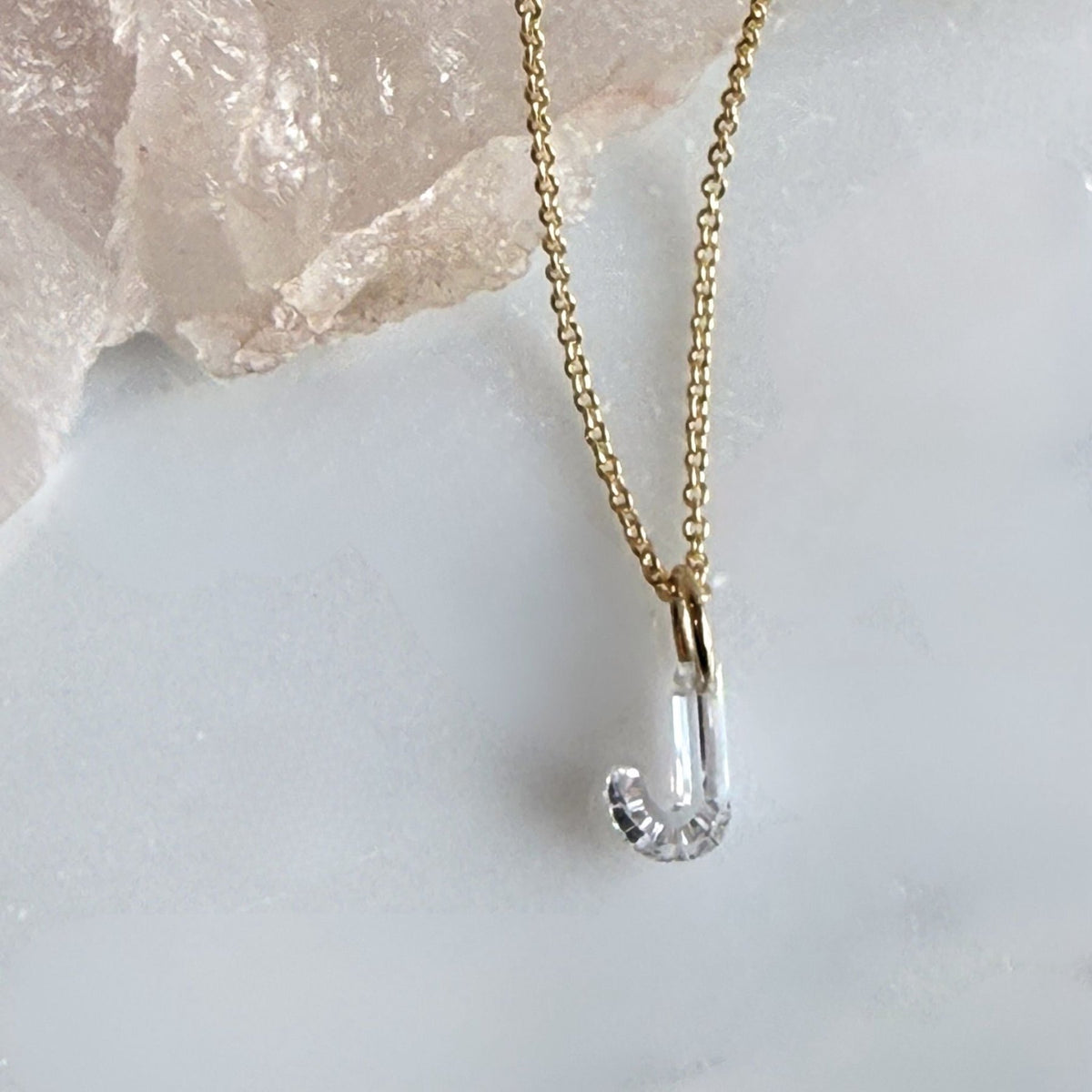 A delicate 14k yellow gold chain necklace with a floating lab created diamond "J" initial charm resting on a light marble surface next to a pink crystal cluster, by Camille Jewelry
