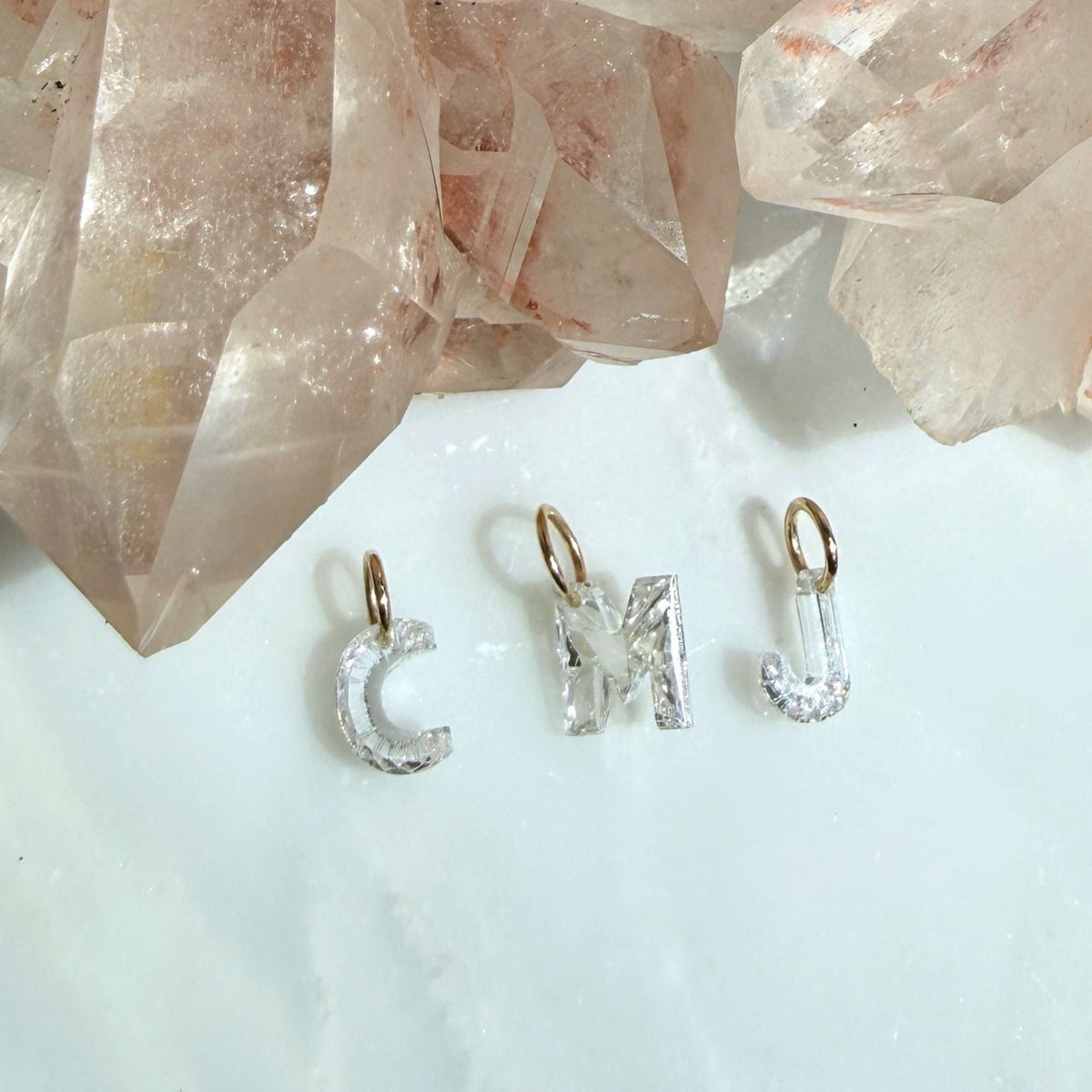 Three lab created floating diamond letter initial charms spelling 'CMJ' with 14k yellow gold jump rings, displayed on a light surface with rose quartz crystals in the background, by camille Jewelry