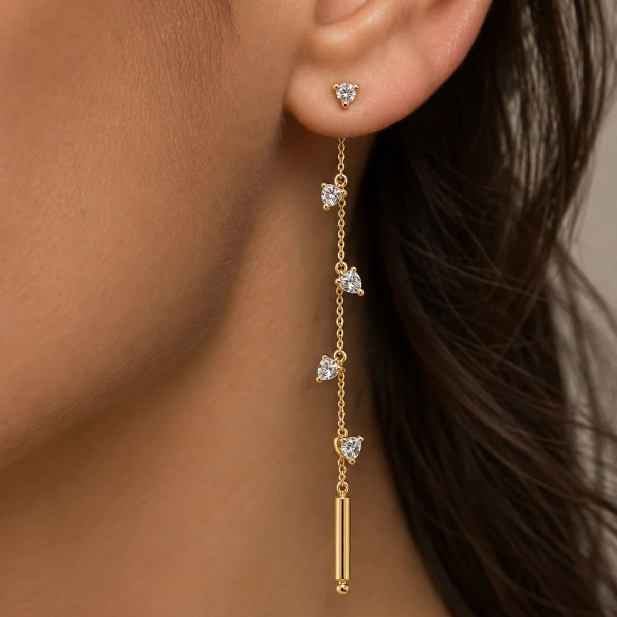 A close-up of a model's ear wearing a gold dangle linear earring with trillion shaped cubic zirconia charms and a gold tube at the end by Camille Jewelry.