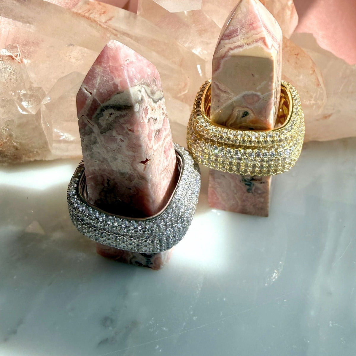 Two square rings displayed on pink stone spires, set against a background of clear quartz crystals. The rings featured are rhodium plated sterling silver  and gold plated silver band paved with clear stones, by Camille Jewelry. 