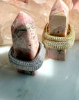 Two square rings displayed on pink stone spires, set against a background of clear quartz crystals. The rings featured are rhodium plated sterling silver  and gold plated silver band paved with clear stones, by Camille Jewelry. 