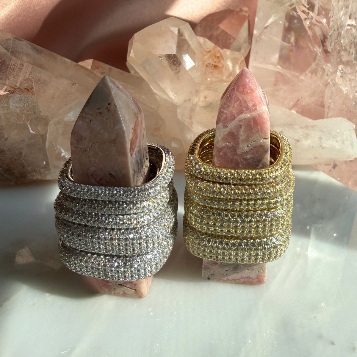 Two square ring styles displayed on pink stone, set against a background of clear quartz crystals. One ring is rhodium plated silver with pavé-set clear stones, and the other is gold plated silver with pavé-set stones by Camille Jewelry
