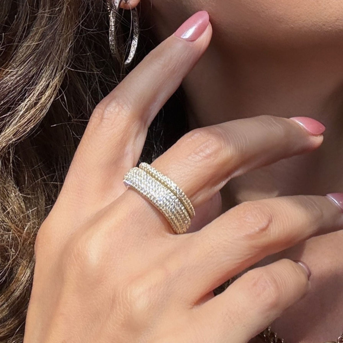 A close-up of a woman's hand wearing a stack of two gold plated sterling silver square pave rings, with pink manicured nails and a silver hoop earring visible, by Camille Jewelry. 