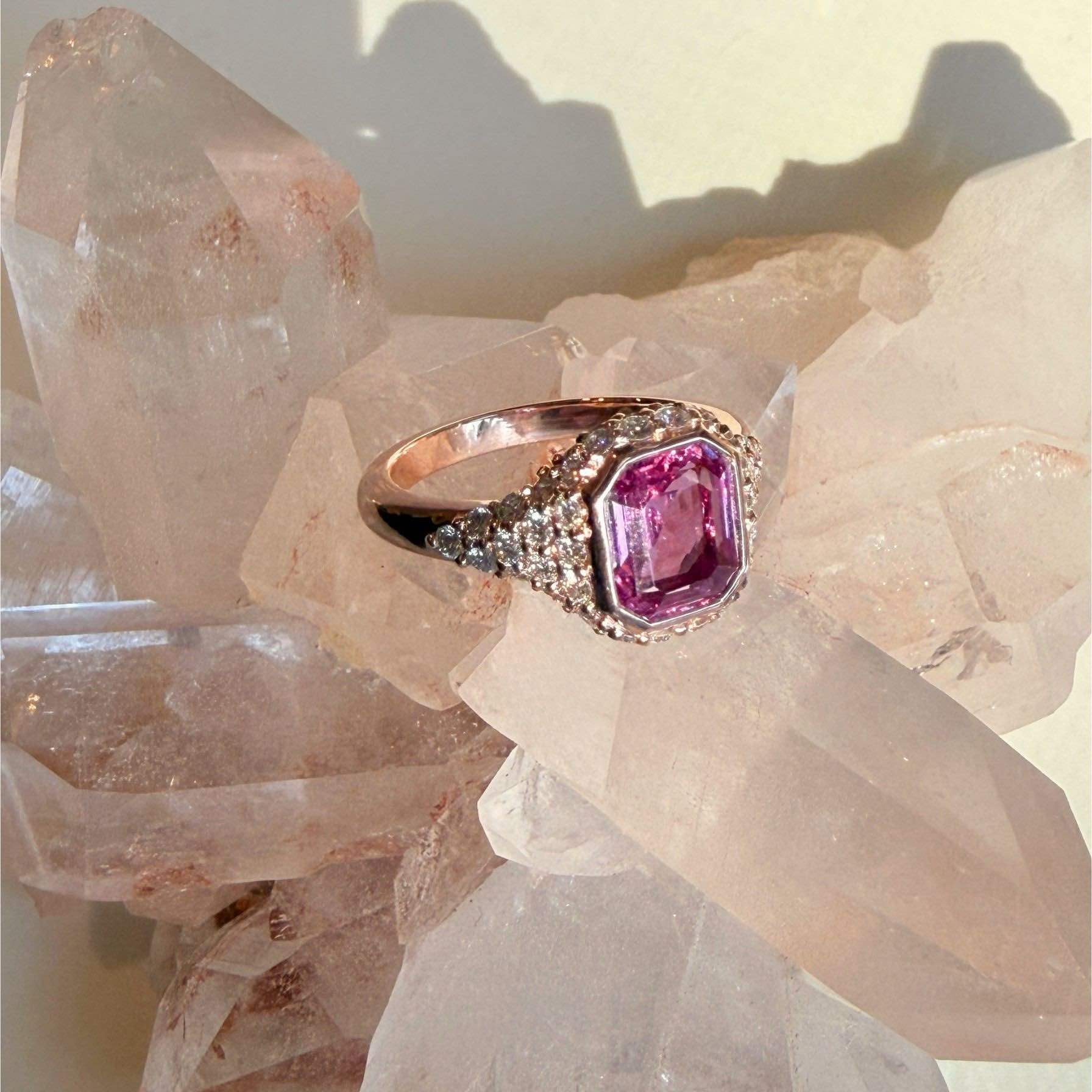 A 14k rose gold ring with a large emerald-cut pink gemstone and pavé-set clear stones, displayed on a cluster of rose quartz crystals by Camille Jewelry