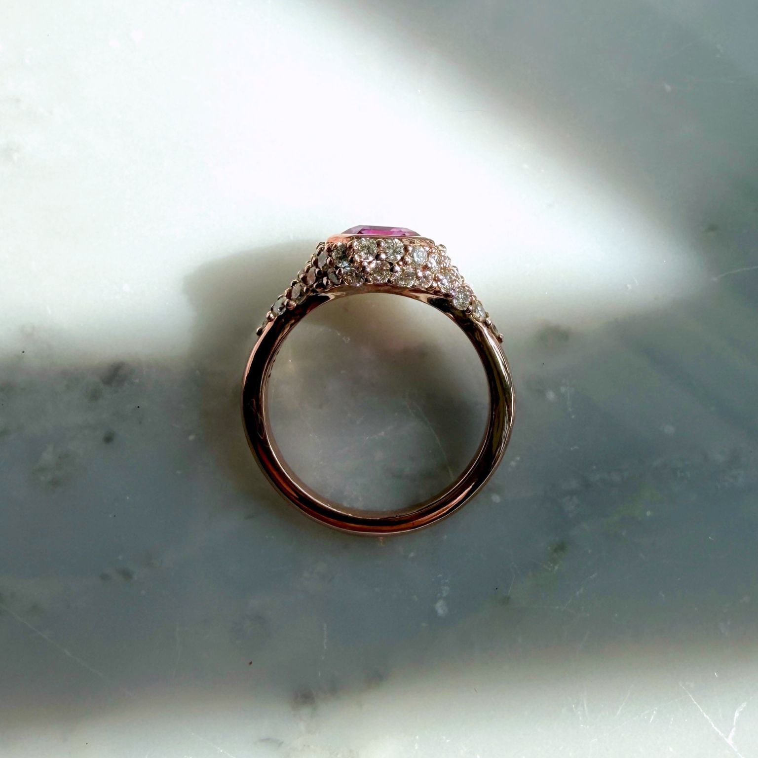A 14k rose gold ring with a pink sapphire gemstone and pave natural diamonds, resting on a light gray marble surface with dramatic shadows by Camille Jewelry.