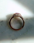 A 14k rose gold ring with a pink sapphire gemstone and pave natural diamonds, resting on a light gray marble surface with dramatic shadows by Camille Jewelry.
