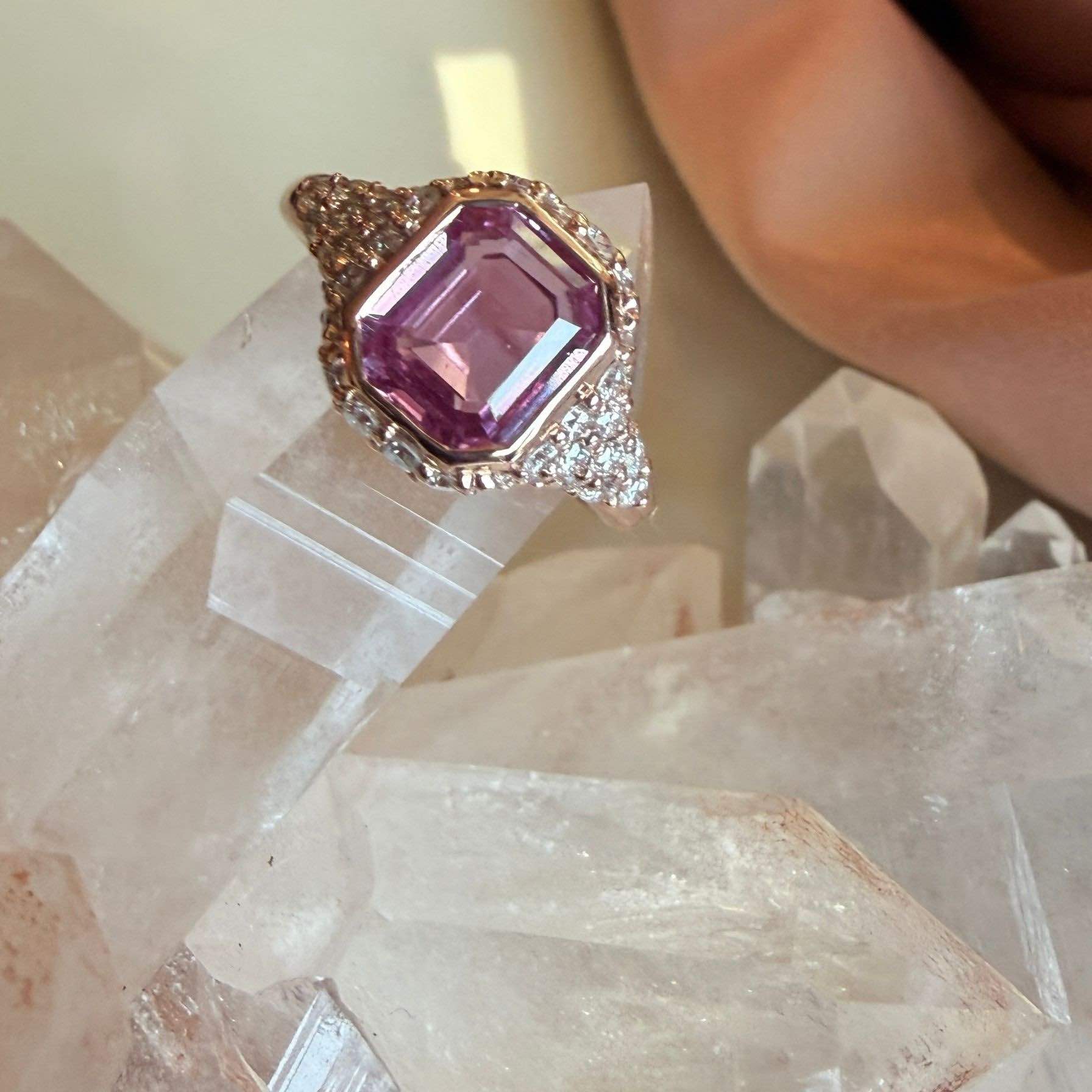 A close-up of a pink sapphire ring with a large emerald-cut gemstone in 14k rose gold setting, accented with pave natural diamonds on a quartz display. 