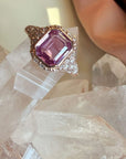 A close-up of a pink sapphire ring with a large emerald-cut gemstone in 14k rose gold setting, accented with pave natural diamonds on a quartz display. 