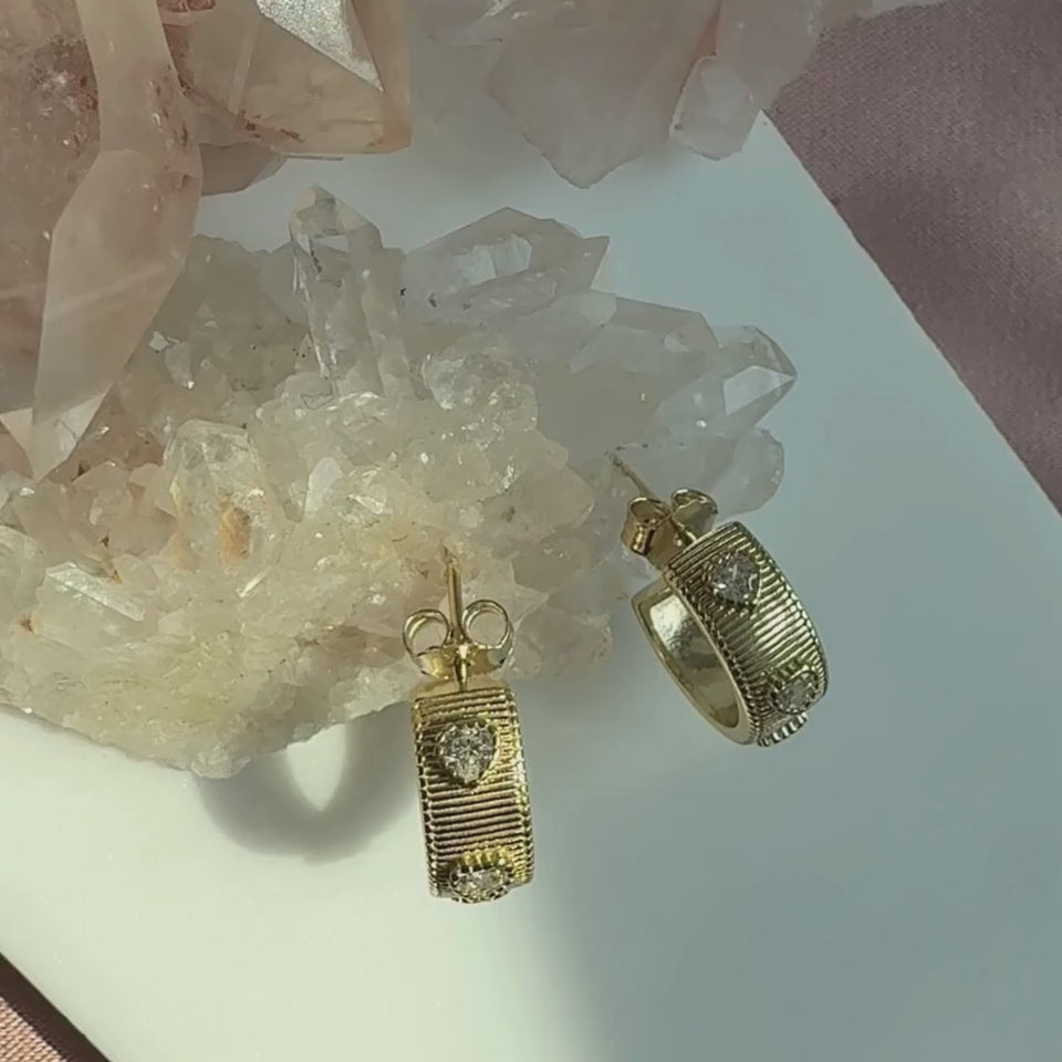 A pair of gold plated sterling silver textured ribbed hoop earrings with heart shaped cubic zirconia stone, displayed on a white surface with crystal clusters in the background by Camille Jewelry. 