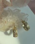 A pair of gold plated sterling silver textured ribbed hoop earrings with heart shaped cubic zirconia stone, displayed on a white surface with crystal clusters in the background by Camille Jewelry. 