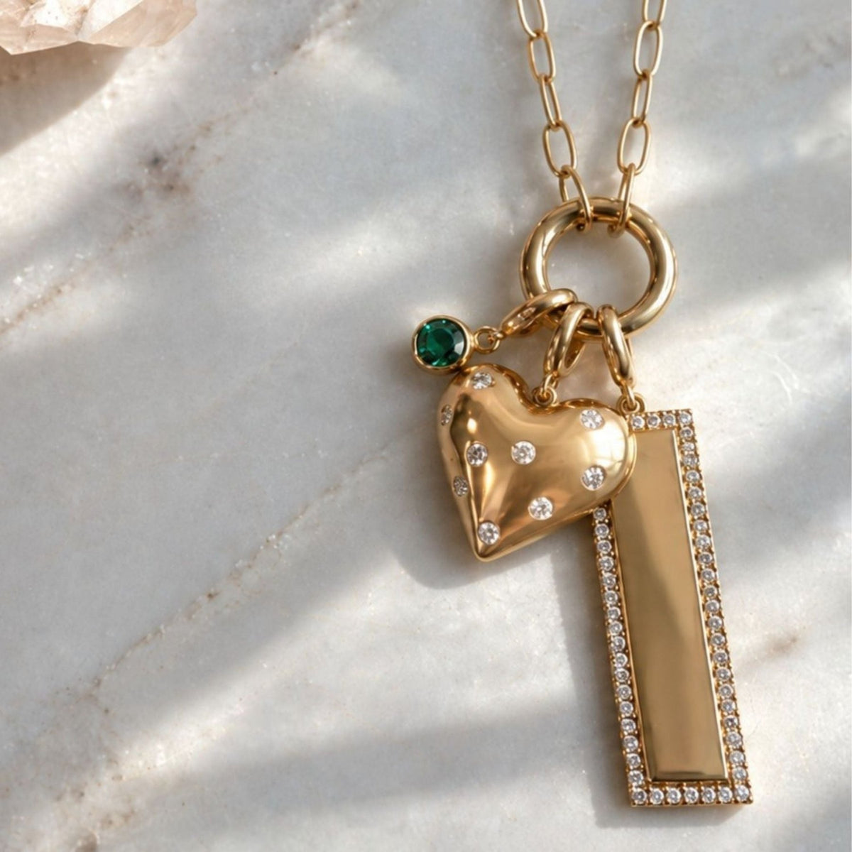 A 14k yellow gold necklace with a multi-charm pendant featuring a puffy heart-shaped charm, a rectangular bar charm, and a small emerald green gemstone accent, displayed on a light marble surface, by Camille Jewelry