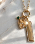 A 14k yellow gold necklace with a multi-charm pendant featuring a puffy heart-shaped charm, a rectangular bar charm, and a small emerald green gemstone accent, displayed on a light marble surface, by Camille Jewelry