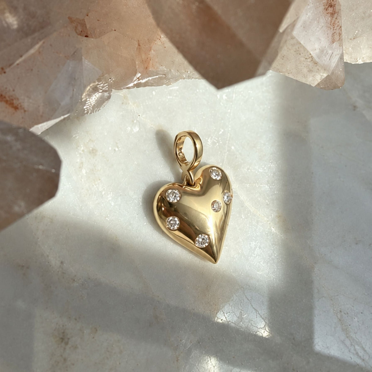 A polished 14k yellow gold puffy heart charm with natural diamonds, displayed on a light marble surface with crystal clusters in the background.