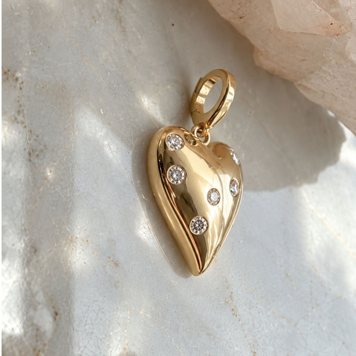 A polished 14k solid yellow gold puffy heart with natural diamonds, photographed on a light marble surface with a soft-focus crystal in the background, by Camille Jewelry. 