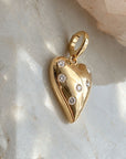 A polished 14k solid yellow gold puffy heart with natural diamonds, photographed on a light marble surface with a soft-focus crystal in the background, by Camille Jewelry. 