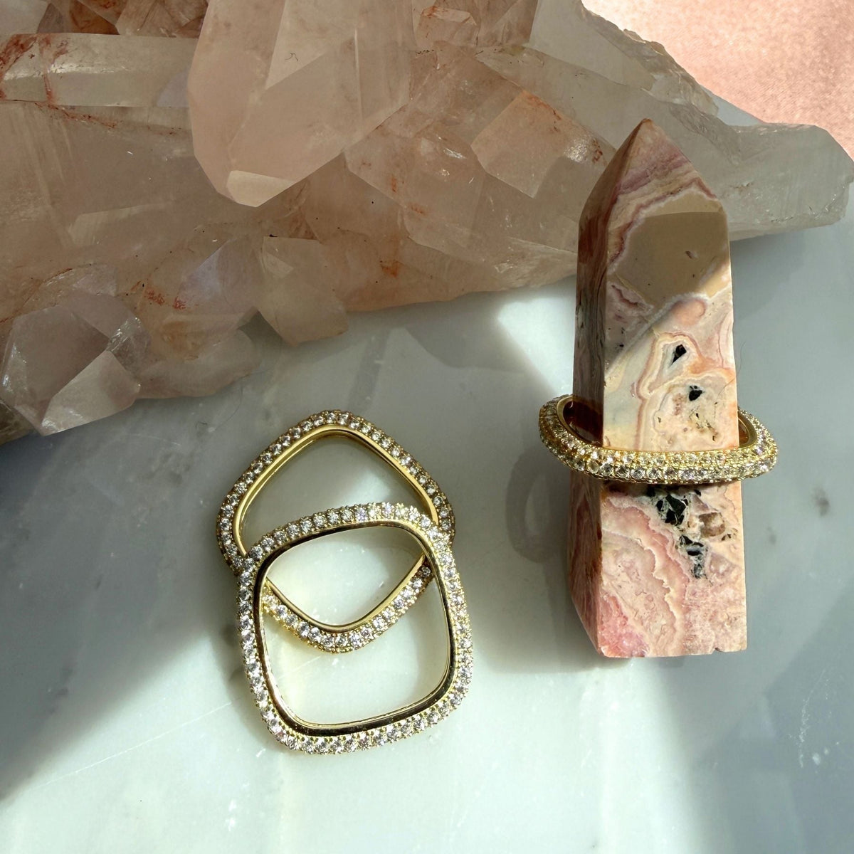 Pave gold plated sterling silver square rings, displayed on a light marble surface with a pink stone agate tower in the background, Camille Jewelry. 