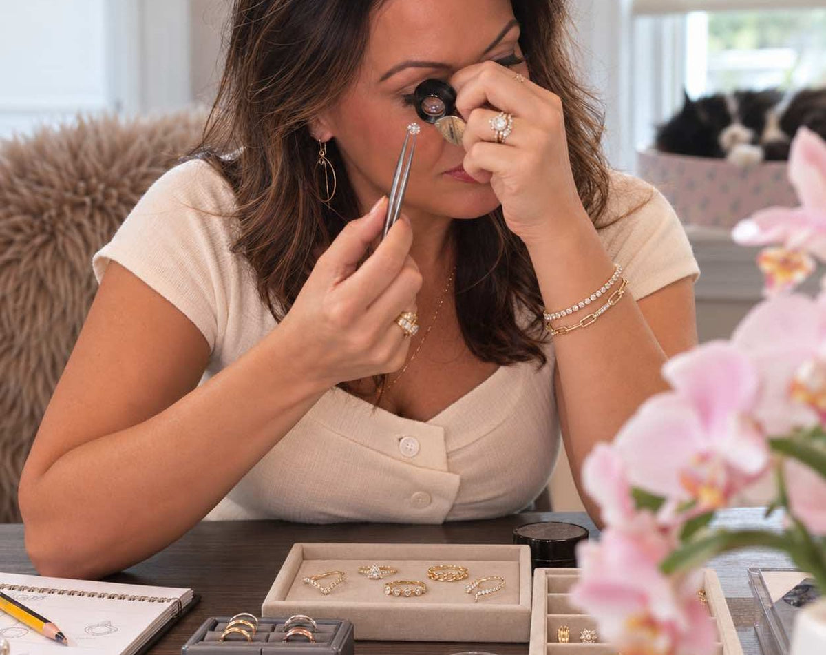 Camille examines small jewelry pieces under magnification at a desk, surrounded by trays of rings, earrings, and gemstones, with pink orchids in the foreground for custom jewelry order at Camille Jewelry.